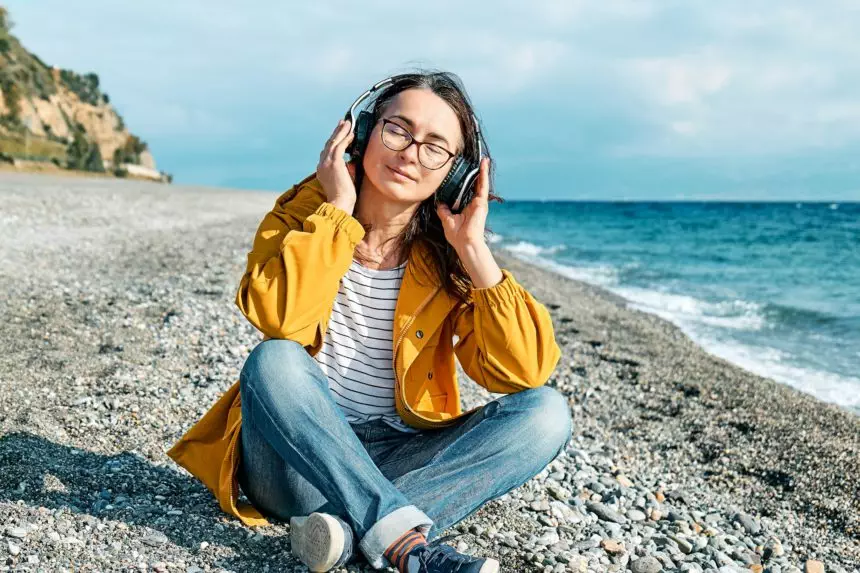 Woman with closed eyes listening music or podcast in headphones while sits on winter beach.